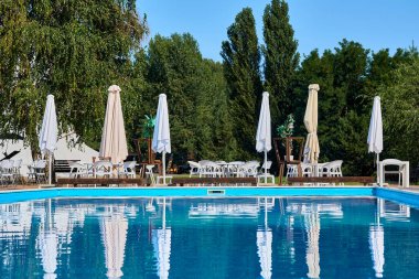 Yeşil doğa üzerinde şemsiyeler ve güneşlikler olan bir dinlenme tesisinde yaz havuzu.