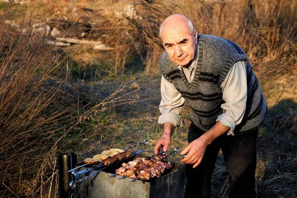Elderly cheerful man pensioner cooking meat on the grill outdoors in nature