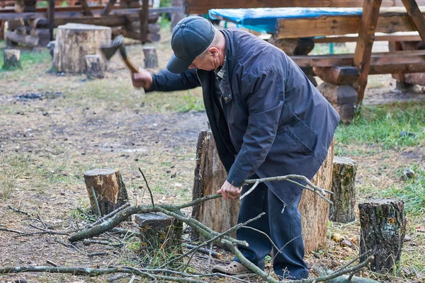 Yaşlı emekli bir adam baltayla odun kesiyor, ateşte ızgara yemek pişiriyor.