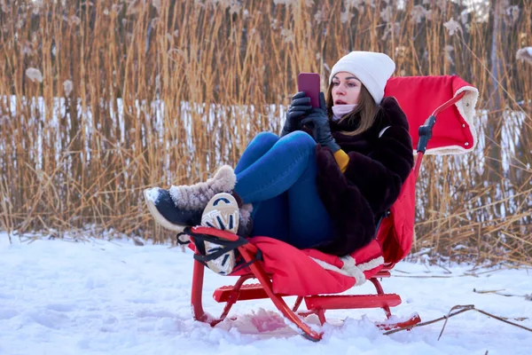 Winter. Pretty woman with mobile phone sits on a red sleigh with a plaid,reeds
