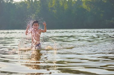 Küçük bir kız yaz nehrinde yıkanıyor, su sıçramalarıyla oynuyor.                               