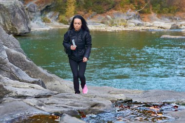 Siyah ceketli genç bir kadın sonbahar nehrinin kayalık kıyısında yürüyor.