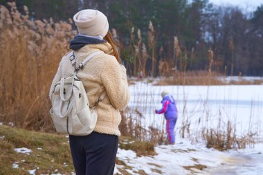 Anne ve kızı parkta, kışın donmuş gölün kıyısında yürüyorlar.