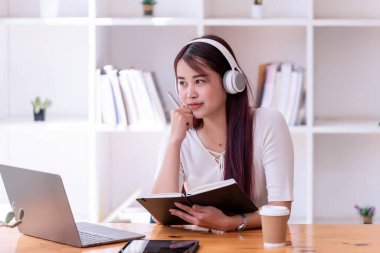 Asyalı kadınlar kulaklık takıyor müzik dinliyor, rahatlıyor ve notlar yazıyor. İnternetten öğrenen güzel bir kadın. Kulaklık takar ve çalışır..