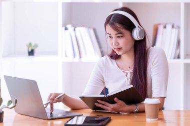 Asyalı kadınlar kulaklık takıyor müzik dinliyor, rahatlıyor ve notlar yazıyor. İnternetten öğrenen güzel bir kadın. Kulaklık takar ve çalışır..