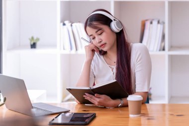 Asyalı kadınlar kulaklık takıyor müzik dinliyor, rahatlıyor ve notlar yazıyor. İnternetten öğrenen güzel bir kadın. Kulaklık takar ve çalışır..