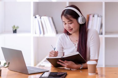 Asyalı kadınlar kulaklık takıyor müzik dinliyor, rahatlıyor ve notlar yazıyor. İnternetten öğrenen güzel bir kadın. Kulaklık takar ve çalışır..