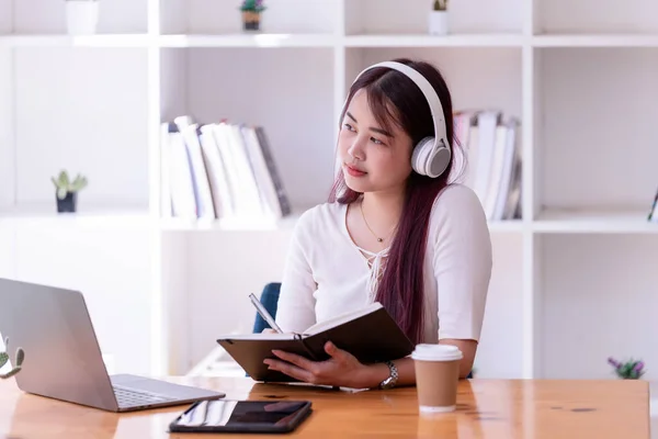アジアの女性はヘッドフォンを着用し 音楽をリラックスしてノートを書きます きれいな女性がオンラインで学ぶ 彼女はヘッドフォンと作品をつけて ロイヤリティフリーのストック写真