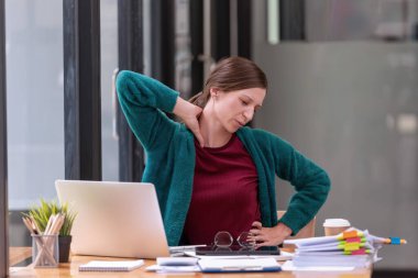 Genç işkadını ofis çalışanı tembelce esneyerek ofis masasının çalışma yerindeki acıyı hafifletiyor, kendini çok yorgun hissediyor. Ofis sendromu.