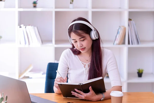 Asian women wear headphones listen to music relax and write notes. Pretty woman learning online. She puts on headphones and works.