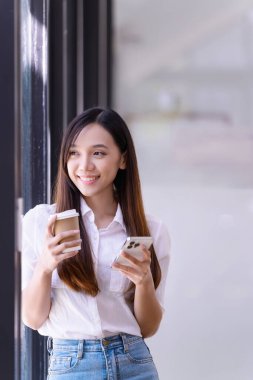 Elinde bir fincan kahve tutan genç bir kadın dinlendirici standı ve ofisteki telefona bakıyor. İş gündelik konsepti.