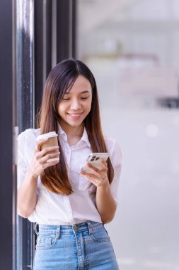 Elinde bir fincan kahve tutan genç bir kadın dinlendirici standı ve ofisteki telefona bakıyor. İş gündelik konsepti.