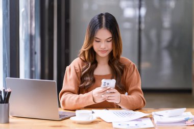 Genç bir kadın ofiste çalışma masasında otururken akıllı telefon kullanmış. İş gündelik konsepti.