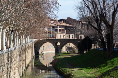 Çevresindeki mimari ve yeşillik ile vurgulanan Vic 'in pitoresk şehrindeki eski bir taş köprünün altından sakin bir nehir akıyor.