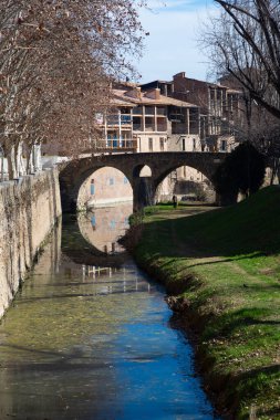 Çevresindeki mimari ve yeşillik ile vurgulanan Vic 'in pitoresk şehrindeki eski bir taş köprünün altından sakin bir nehir akıyor.