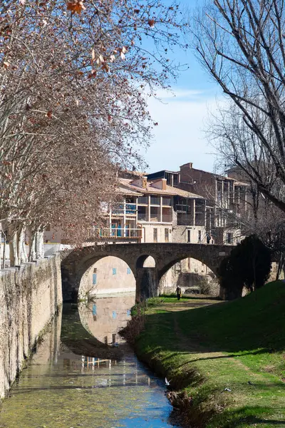 Çevresindeki mimari ve yeşillik ile vurgulanan Vic 'in pitoresk şehrindeki eski bir taş köprünün altından sakin bir nehir akıyor.