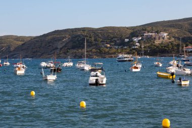 Tekneler huzur içinde yüzüyor, arka planda dağlık bir kıyı şeridi ve tuhaf bir köyü olan bir limana yanaşmış durumdalar.