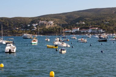 Tekneler huzur içinde yüzüyor, arka planda dağlık bir kıyı şeridi ve tuhaf bir köyü olan bir limana yanaşmış durumdalar.