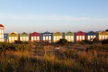 Renkli plaj kulübelerinin sırası sahilde duruyor. Arka planda okyanus var.