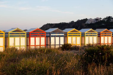 Renkli plaj kulübelerinin sırası sahilde duruyor. Arka planda okyanus var.