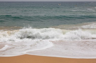 Turkuaz deniz suyu, arka planda tekneler olan kumlu bir sahilde kırılıyor.