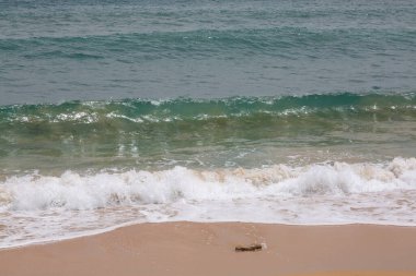 Turkuaz deniz suyu, arka planda tekneler olan kumlu bir sahilde kırılıyor.