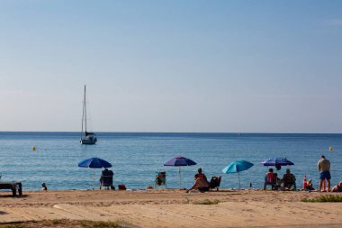 Turistler güneşli bir sahilde güneşli bir günün tadını çıkarıyorlar. Şemsiyelerin altında sakin mavi sularda görünen bir yelkenliyle dinleniyorlar.