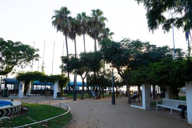 İnsanlar gün batımını ağaçlı, banklı ve sokak lambalı bir parkta severler.