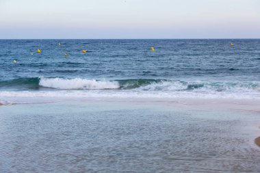 Sakin bir okyanusta kumlu bir sahilde kıyıya vuran küçük dalgalar ve sarı şamandıralar.