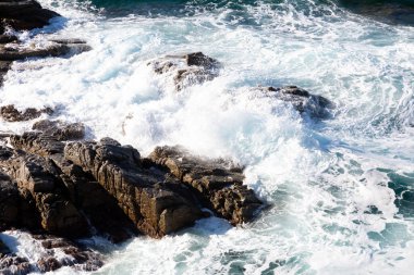 Beyaz okyanus dalgaları güneşli bir günde karanlık kayalara çarpıyor.