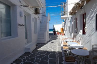 Boş beyaz masalar ve sandalyeler Yunan adasının pitoresk geçidinde duruyor, güneş ışığıyla yıkanmış.