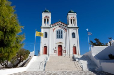 Geleneksel beyaz kilise, kırmızı kapılar, Bizans bayrağı ve derin mavi gökyüzünün altında rüzgarda dalgalanan Yunan bayrağı.