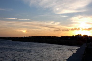 Altın gün batımı ince bulutları aydınlatıyor ve kıyı şeridi siluetiyle sakin denizi.