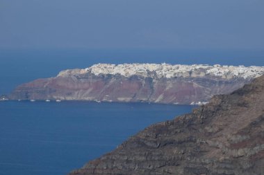 Santorini Adası 'ndaki beyaz imerovigli kasabası gün ışığında mavi deniz ve önplandaki karanlık volkanik kayalarla renkli kaldera uçurumlarının üzerinde yükseliyor.