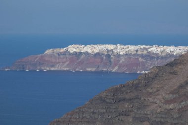 Santorini Adası 'ndaki beyaz imerovigli kasabası gün ışığında mavi deniz ve önplandaki karanlık volkanik kayalarla renkli kaldera uçurumlarının üzerinde yükseliyor.