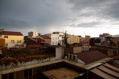 Terasları ve çatıları olan yerleşim yerleri bulutlu bir gökyüzü altında kentsel bir manzara oluşturarak, havanın değişmesinin an meselesi olduğunu ima ediyor.