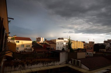 Terasları ve çatıları olan yerleşim yerleri bulutlu bir gökyüzü altında kentsel bir manzara oluşturarak, havanın değişmesinin an meselesi olduğunu ima ediyor.