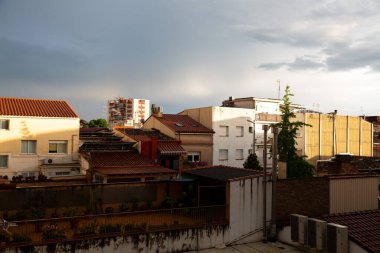 Terasları ve çatıları olan yerleşim yerleri bulutlu bir gökyüzü altında kentsel bir manzara oluşturarak, havanın değişmesinin an meselesi olduğunu ima ediyor.