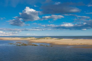 Sakin kıyı manzarası bulutlu bir gökyüzünün altında kumlu bir plajda kıyıya vuran yumuşak dalgaların oluşturduğu sakin bir atmosfer yaratıyor.
