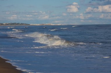 Sakin kıyı manzarası bulutlu bir gökyüzünün altında kumlu bir plajda kıyıya vuran yumuşak dalgaların oluşturduğu sakin bir atmosfer yaratıyor.