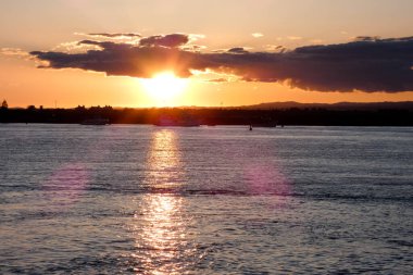 Gün batımında turuncu ve pembe bulutlarla sakin okyanus suyunun üzerinde uçan kuş.