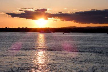 Gün batımında turuncu ve pembe bulutlarla sakin okyanus suyunun üzerinde uçan kuş.