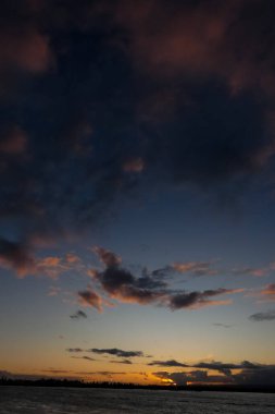 Gün batımında turuncu ve pembe bulutlarla sakin okyanus suyunun üzerinde uçan kuş.