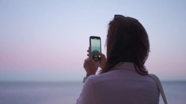 Kadın gezgin gün batımında akıllı telefon kamerasıyla denizin fotoğrafını çekiyor. Yetişkin bir kadının telefon kamerasıyla video deniz manzarası çekerken arka plan portre modu yavaş çekim