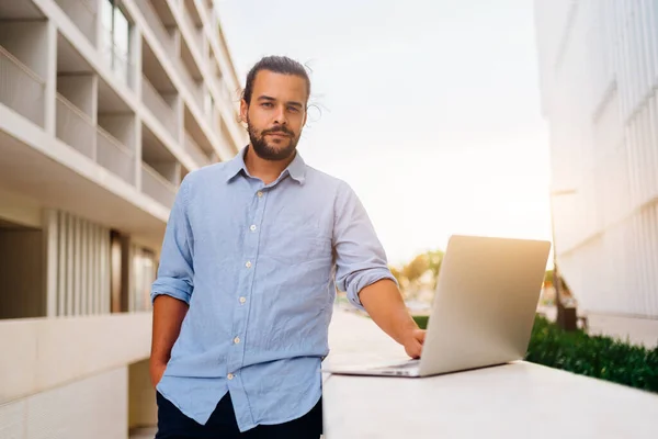 Hispanic businessman standing outside with laptop working remote distant job outdoors near office company building. Stylish unshaved male business man with long hair dressed use laptop