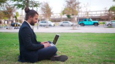 Handsome hispanic male businessman sitting lotus position in public park use laptop. Remote work concept. Stylish freelancer in suit relax in park summer day working on computer. Technology. Handheld.