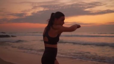 Gün batımında deniz kıyısındaki plajda boks yapan bir sporcu. Midilli kuyruklu çekici bir kadın elleriyle vuruyor, spor yapıyor, formda, orta yaşlı bayan kickboksör. 