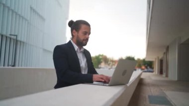 Hispanic businessman standing outside with laptop working remote distant job outdoors near office company building. Stylish unshaved male business man with long hair dressed business suit use laptop