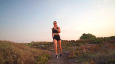 Doğada koşu yapan bir kadın kırsal kesim manzarası izliyor. Sağlıklı koşu ve açık hava egzersizlerini, ağır çekim el kameralarını hesaba katın. Beyaz, orta yaşlı kadın koşucu gün batımında koşuyor.