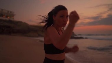 Gün batımında deniz kıyısındaki plajda boks yapan bir sporcu. Midilli kuyruklu çekici bir kadın elleriyle vuruyor, spor yapıyor, formda orta yaşlı bayan kickboksör yakın plan.